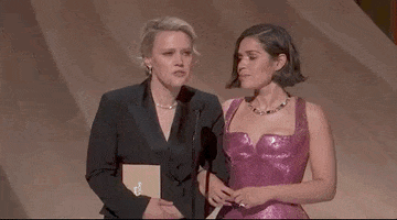 Oscars 2024 GIF. Kate McKinnon and America Ferrera stand on stage and McKinnon leans into the microphone to inquire with utter seriousness, "To whom have I been sending my tasteful nudes?" The camera pans to Steven Spielberg, who slowly turns to the camera and points exaggeratedly at himself, with a save-me expression on his face. 
