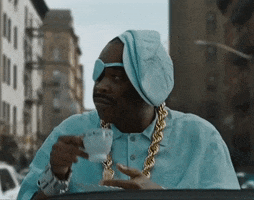 Music video gif. From the video for French Montana's "No Stylist," Slick Rick wears a light blue shirt, hat, and eyepatch, and sips tea while sitting up in a convertible driving through a city.