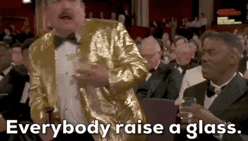 Oscars 2024 GIF. Guillermo stands up from his seat and immediately turns his back on the camera, addressing the audience and holding his margarita up. Colman Domingo sits next to him and starts laughing.