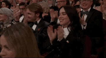 Oscars 2024 GIF. Billie Eilish, seated at the Oscars, applauds energetically, throwing in a whoop and cheer.