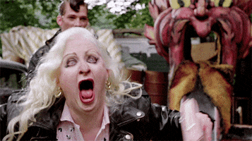 Video gif. An older woman wearing a leather jacket with platinum blonde hair tries to scare us by yelling and flailing her arms. In the background is a slide that runs through the mouth of an ornamental cartoon devil head.