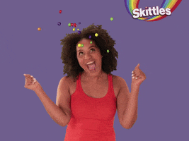 Video gif. A woman with curly hair and a sleeveless orange shirt looks upward, rocking back and forth in a very excited dance as Skittles float in the air above her.