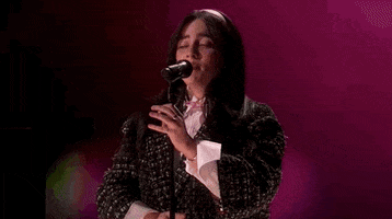 Oscars 2024 GIF. Billie Eilish performing "What Was I Made For" on stage at the Oscars. Eilish holds the stand of the microphone gently and she shakes her head and hair out as she hits a high note. The background has bokeh colors of pink and green.
