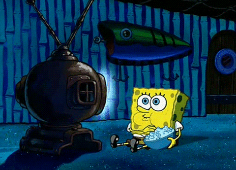 TV gif. SpongeBob sits on the floor in a dark room with his face lit up by a TV screen. He holds a bowl of popcorn in his arms. His eyes are glued to the TV screen, completely captivated by it, as he munches delightfully on the popcorn. 