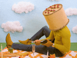 Video gif. A man with a birthday cake for a head, sits on a picnic blanket with a fancy, romantic meal spread out for us. The backdrop is construction paper grass, sky, and cotton-ball clouds. He opens a bottle of champagne which sprays everywhere before holding his hand out in presentation. Text, "Happy anniversary."