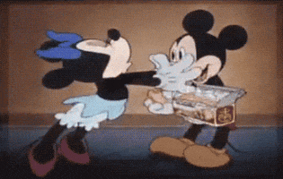 Disney gif. Minnie lunges forward and grabs Mickey around his body, planting kisses all over his face. He looks happy as he tosses a box he's holding to the side.