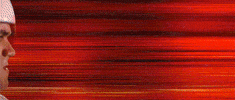 Movie gif. From the left side of the frame, the helmeted head of Emile Hirsch as Speed Racer emerges, zooming fast through a blurry reddish background.