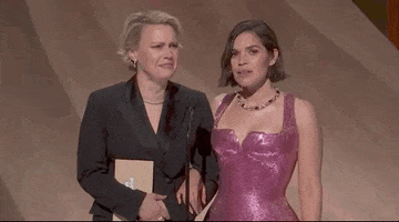 Oscars 2024 GIF. Kate McKinnon and America Ferrera stand with linked arms at the microphone and McKinnon pulls a face at something Ferrera has said. She scrunches her face up and juts her chin out incredulously as she asks, "What do you mean?"