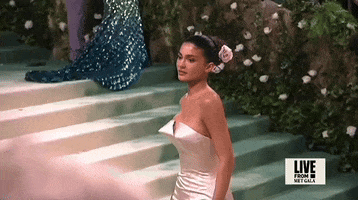 Met Gala 2024 gif. Slow motion close up of the top of Kylie Jenner's Oscar de la Renta champagne-colored strapless column gown featuring a pointed bust. She also wears a pale pink rose in a smooth mid bun.