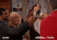 Parks and Recreation gif. Amy Poehler, Aziz Ansari, Nick Offerman, and Jim O'Heir as Leslie, Tom, Ron, and Jerry cheerfully clink champagne glasses with each other and other colleagues.