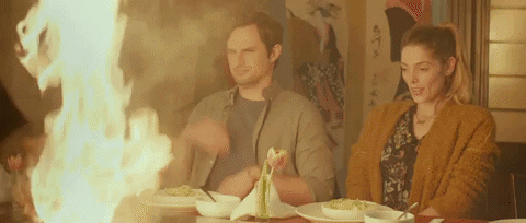 Movie gif. Hibachi grill flame billows in front of Andrew J. West as Walt in Antiquities, who covers his face protectively and nervously while Ashley Greene as Ellie smiles and laughs.