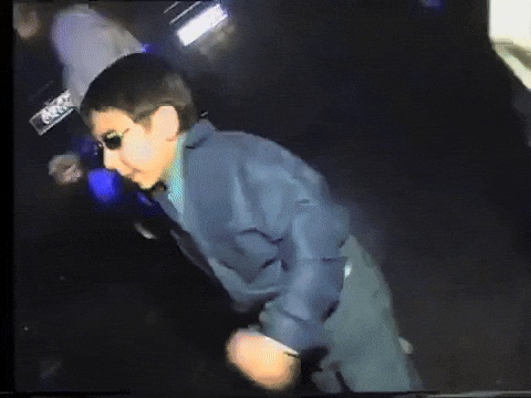 Video gif. Young boy in sunglasses and adult attire, dancing and boogeying amongst adults in a dark nightclub.