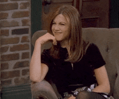 Friends gif. Jennifer Aniston as Rachel sitting down, squinting and wrinkling her nose, and giving a flirty wave.