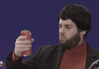Video gif. A man with a bad black wig on stares at a soda can that he holds out and confidently crushes with his hand. 
