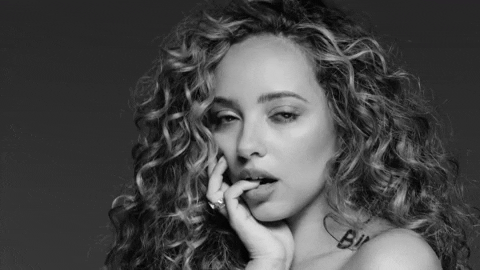 Celebrity gif. Black-and-white video of Jade Thirlwall cupping her face with her hand, her little finger in her mouth, shrugging and giving a flirty eyeroll.