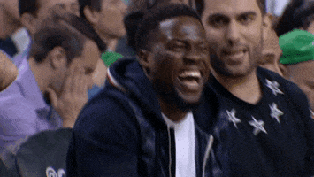 Sports gif. Kevin Hart sits courtside at a basketball game in a dark blue coat, laughing at a comment from the stubbly man next to him.