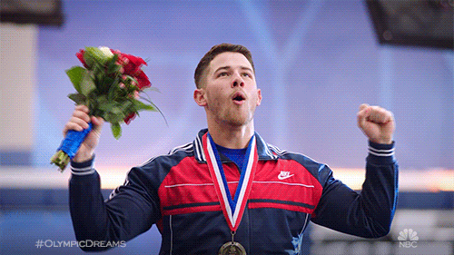 TV gif. Nick Jonas, dressed as an Olympian, wears a medal around his neck and raises his arms to the sky in victory, his mouth open in a "wooo!" shape. He's obviously just won an Olympic medal for being the cutest Jonas Brother.