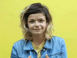 Video gif. Person looks at us with a small smile and brings their hands up in a prayer position up to their mouth.
