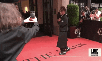 Met Gala 2024 gif. Jaden Smith walks into the entrance of The Mark holding up two peace signs for camera. He's wearing a loose fitting boxy half zipped black coat over a white collared shirt and black tie. This is paired with a black towel skirt that falls below the knees over black trousers and black and white sneakers.