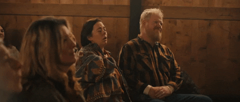 Movie gif. Olivia Colman as Hope and Jim Gaffigan as Zeke in "Them That Follow" sit amongst churchgoers; Hope raises her hand up with eyes closed and Zeke puts his hand on his knee and leans back.