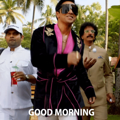 Celebrity gif. Trailed by his entourage, Varun Dhawan struts happily, wearing a pink and black robe and giant sunglasses. Text, “Good Morning.”