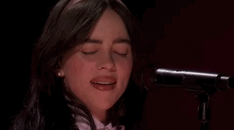 Oscars 2024 GIF. Close up shot of Billie Eilish performing "What Was I Made For" on stage at the Oscars. Eilish sings the last word of the song and pulls away from the microphone, looking at Finneas and grinning. 