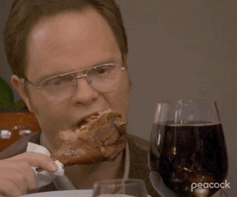 The Office gif. Rainn Wilson as Dwight gnaws on a turkey leg with an enormous glass of wine, at a table with coworkers.