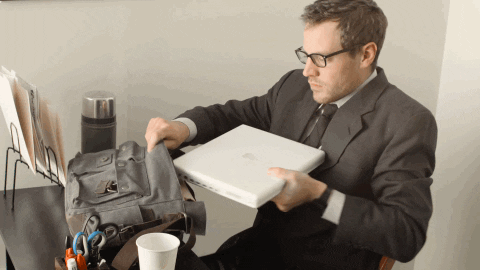 Video gif. Man wearing a suit and sitting at a desk struggles to stuff a MacBook into a messenger bag, from the music video for "Worth" by Clique.