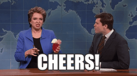 SNL gif. On Weekend Update, Maya Rudolph raises a cocktail and says "cheers!" at news desk while Colin Jost watches her and then looks back at us.