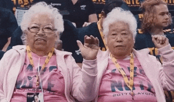 Sports gif. Cute centenarian twin sisters, sitting in the stands at a sports event, wave at us placidly. 