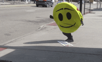 Video gif. A yellow smiley faced mascot slowly falls to their knees to try to pick up the sign that’s on the sidewalk. The mascot loses their balance and falls backwards. 