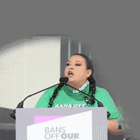Video gif. Woman wearing a bright green t-shirt speaks emphatically at a podium above a sign that reads, "Bans off our bodies." She says, "The day we warned about is here, the end of Roe and Casey are in plain sight, and we will not go back."