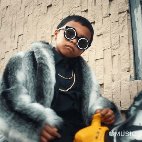 Music video gif. In Anderson Paak's video for Bubblin, a little boy wearing a gold chain and a furry jacket stares us down while riding a coin-operated horse.