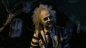 Movie gif. Michael Keaton as Beetlejuice sits in a graveyard while the lightning flashes. He holds his arms out and smiles as he says, “It’s showtime.” 
