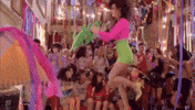 Music Video gif. Katy Perry is dancing on a table during her song, Last Friday Night. She shuffles as she moves her hands back and forth and the party around her goes wild.