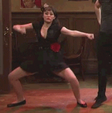 TV gif. A young woman, wearing a black dress with a red flower ornament on her hip and a feathery hair decoration, does a provocative modern dance. Next to her, a man dressed in black does an Irish step dance.