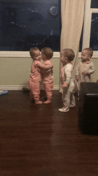 Video gif. Four babies all stand up. Two babies in pink hug each other while the other two awkwardly stand and wait their turn. One of the babies in white taps a baby in pink and they hug. The four babies then go around, hugging each other and smiling with excitement as they pat each other’s backs. 