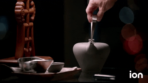 Video gif. Hand pours clear liquid out of a sleek gray teapot into a small gray cup. An intricate piece of smooth woodwork rests behind the cup, and the background is dark with hazy glints of red and blue lights.