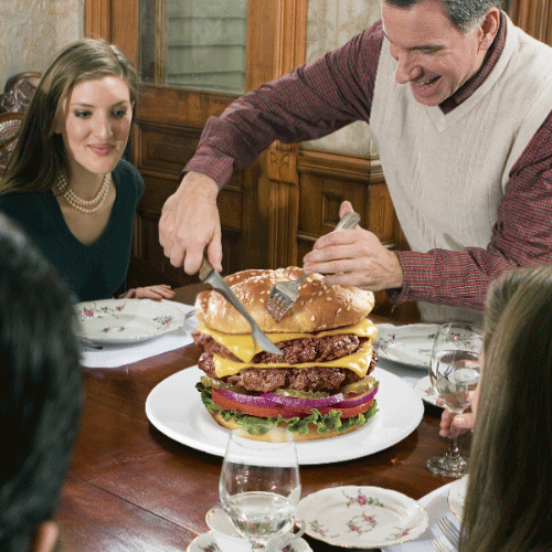 Digital art gif. Animated from still images, a family is gathered around a dinner table, and a smiling man in a sweater vest is carving...a giant double cheeseburger.