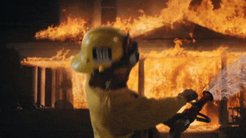 Celebrity gif. BRS Kash is dressed in a firefighter’s suit and stands in front of a house on fire. He leans back as he hoses down the house, bouncing back and forth as he raps. 