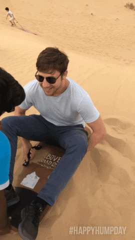 Video gif. A man sits on a small sled and gets pushed down a large desert sand dune backwards.