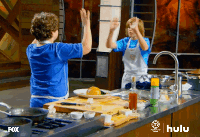 Reality TV gif. Two boys on Masterchef Junior high five with both hands and then look down at the plate they made together.