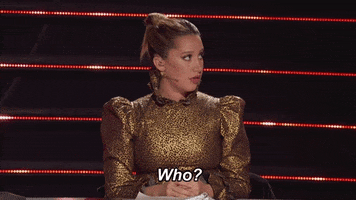 TV gif. A scene from The Masked Dancer. Wearing a shiny gold dress with puffy sleeves and a tall collar, Ashley Tisdale gives a dull, minimal reaction to right of frame. Text, "Who?"