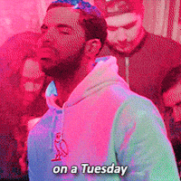 Music video gif. Young Drake is rapping in a club and there are people sitting around him, grooving and drinking. He says, "On a Tuesday," while looking at us and pointing. 