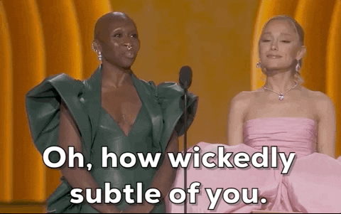 Oscars 2024 GIF. Ariana Grande and Cynthia Ervo stand at the microphone and Grande turns to Ervo with a small smile, saying, "Oh, how wickedly subtle of you." Ervo turns back and says, "I do try."