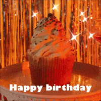 Video gif. A cupcake with frosting piles high sits on a plate in a dimly lit space with metallic streamers twinkling in the background. Text, "Happy birthday."