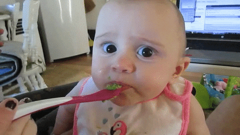 Video gif. Closeup of a baby being fed a spoonful of food, then pursing their lips and furrowing their brow in disgust.