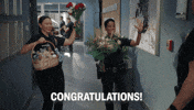 TV gif. On The Rookie, an excited Mekia Cox as Nyla offers a bouquet while Melissa O’Neil as Lucy offers a gift basket and roses to a coworker. Text, “Congratulations!”