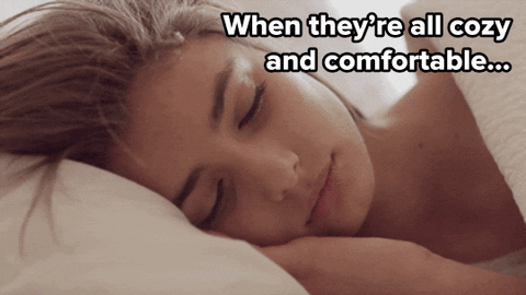 Video gif. A woman with large eyelashes lying in bed opens her eyes to look at us seductively. Text, "When they're all cozy and comfortable..."