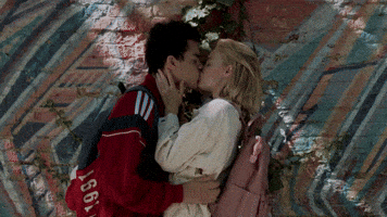 Video gif. Man and a woman are standing in front of a brick wall and the man grabs the woman's butt and kisses her.